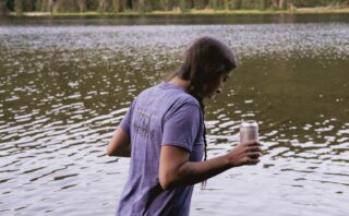 Woman holding a beer next to a mountain lake.