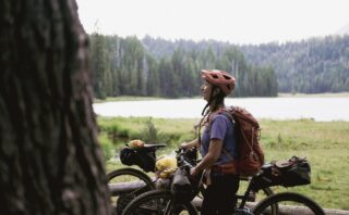 Woman straddling a mountain bike loaded with gear with a mountain lake in the background.