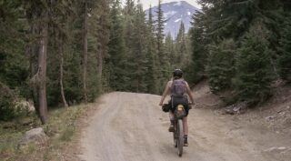 Woman riding a loaded mountain bike on a fire road with a mountain in the distance.